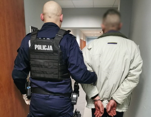 Umundurowany policjant i zatrzymany. Zdjęcie zrobione od tyłu, mężczyzna ma założone kajdanki na ręce z tyłu.