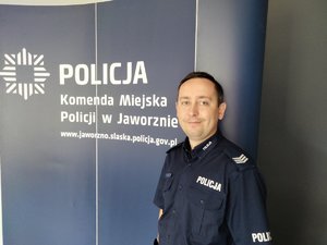 Dzielnicowy w umundurowaniu służbowym na tle ścianki z napisem Komenda Miejska Policji w Jaworznie