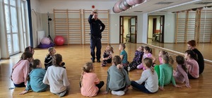 na zdjęciu widoczna siedząca grupa dzieci na sali gimnastycznej, przy której stoi umundurowany policjant trzymający w górze lizaka policyjnego