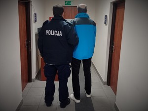 na zdjęciu umundurowany policjant stojący tyłem, trzymający zatrzymanego.