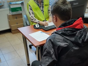 na zdjęciu zatrzymany mężczyzna siedzi przy biurku, a na przeciw policjantka wykonująca z nim czynności