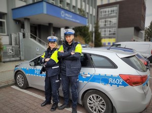 na zdjęciu dwóch umundurowanych policjantów stojących przy radiowozie policyjnym, pod budynkiem Komendy Miejskiej Policji w Jaworznie