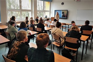 na zdjęciu uczniowie siedzący w ławkach w klasie szkolnej oraz stojąca przed nimi kobieta