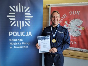 na zdjęciu umundurowany policjant z medalem i dyplomem w ręce, na tle szyldu komendy miejskiej policji w jaworznie i sztandaru jednostki