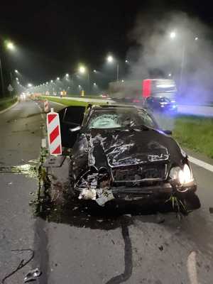 na zdjęciu roztrzaskany samochód stojący na autostradzie w kolorze czarnym, z którego się dymi