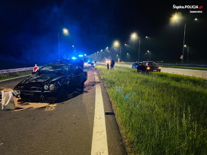 na zdjęciu roztrzaskany samochód stojący na autostradzie w kolorze czarnym, z którego się dymi