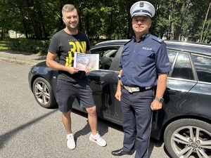 na zdjęciu po prawej stronie umundurowany policjant, po lewej stronie mężczyzna, który trzyma w rękach plakat z napisem &quot;Bezpieczna Prędkość&quot;.