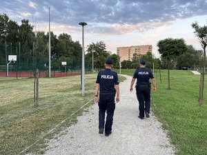 na zdjęciu dwóch umundurowanych policjantów, którzy patrolują park i idą w stronę boiska