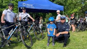 na zdjęciu widoczny jest policyjny namiot profilaktyczny przed którym stoi dwóch umundurowanych policjantów w patrolu rowerowym oraz umundurowany policjant, który kuca przy dziecku