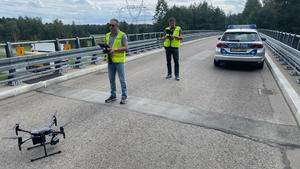 na zdjęciu dwóch umundurowanych policjantów, którzy obsługują drona. W tle radiowóz  policji.