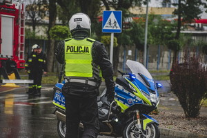 na zdjęciu umundurowany policjant ruchu drogowego tyłem w kamizelce odblaskowej, idzie w stronę motocyklu policyjnego