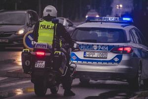 na zdjęciu policjant ruchu drogowego tyłem na policyjnym motocyklu, który stoi na ulicy za radiowozem policyjnym