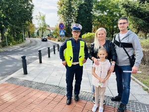 na zdjęciu rodzina z dzieckiem stoi wraz z umundurowanym policjantem przy przejściu dla pieszych, wszyscy trzymają elementy odblaskowe