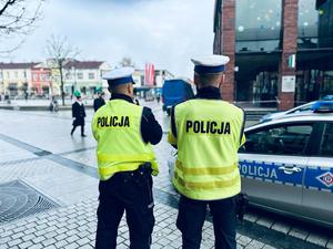 na zdjęciu umundurowani policjanci w kamizelkach odblaskowych stoją przy radiowozie i obserwują Rynek Główny