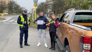 na zdjęciu umundurowany policjant stojący obok mężczyzny trzymającego ulotkę i kobiety trzymającej odblask