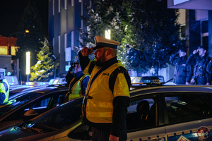 na zdjęciu umundurowany policjant stojący przy radiowozie oddający hołd, za nim stoją również inni umundurowani policjanci oddający hołd