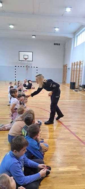 na zdjęciu policjantka na sali gimnastycznej przykłada mikrofon dla jednej z dzieci