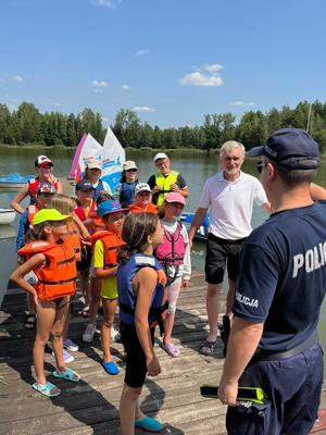 na zdjęciu grupa dzieci oraz starszy mężczyzna, którzy stoją przed umundurowanym policjantem na pomoście