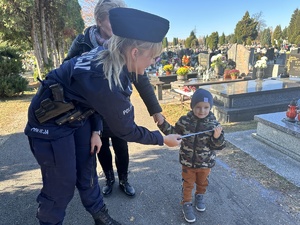 na zdjęciu umundurowana policjantka wręcza opaskę odblaskową dziecku na cmentarzu