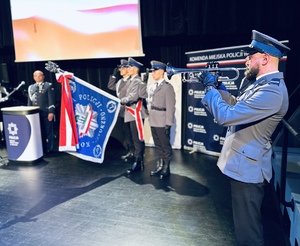 na zdjęciu umundurowany policjant grający na trąbce, w tle umundurowani policjanci stojący na baczność ze sztandarem oddający honor