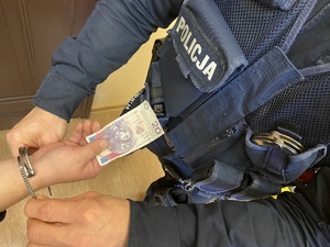 na zdjęciu osoba zatrzymana w kajdankach wystawia banknot w nominale 20zł w stronę umundurowanego policjanta