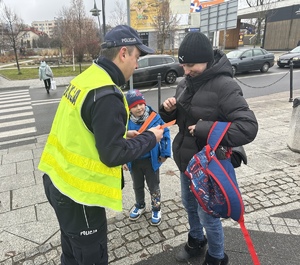 na zdjęciu umundurowany policjant wręcza opaskę odblaskową kobiecie z dzieckiem i plecakiem