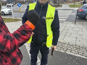 na zdjęciu ręka mężczyzny z założoną opaską odblaskową, a w tle policjant umundurowany