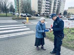 na zdjęciu umundurowany policjant wręcza starszej kobiecie opaskę odblaskową