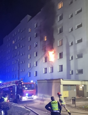 na zdjęciu widoczny jest umundurowany policjant, który zabezpiecza miejsce pożaru, strażak oraz wóz strażacki. Z płonącego mieszkania widoczny jest ogień