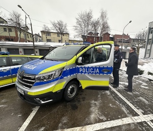 na zdjęciu oznakowany radiowóz furgon, do którego wsiada mężczyzna. Obok stoi umundurowany policjant oraz drugi mężczyzna