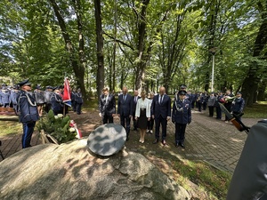 na zdjęciu uczestnicy uroczystości stoją pod obeliskiem i oddają hołd, po prawej stronie umundurowani policjanci, po lewej stronie pododdział policjantów z dowódcą