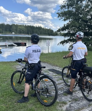 na zdjęciu umundurowany policjant w patrolu rowerowym oraz umundurowany strażnik miejski, którzy zwróceni są w kierunku zalewu Sosina