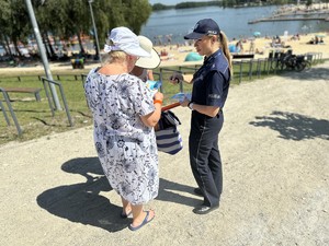 na zdjęciu umundurowana policjantka wręcza książeczkę edukacyjną kobietom odpoczywającym nad akwenem wodnym