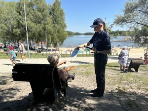 na zdjęciu policjantka w mundurze wręcza książeczkę edukacyjną kobiecie odpoczywającej nad wodą