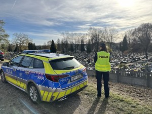 na zdjęciu umundurowana policjantka stoi przy radiowozie i obserwuje cmentarz