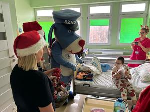 na zdjęciu policyjna maskotka Sznupek wraz z dzieckiem które trzyma prezent i personelem szpitala