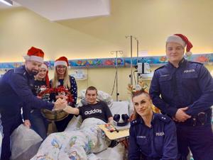 na zdjęciu umundurowani policjanci w czapkach mikołajkowych, jeden z policjantów przybija piątkę dziecku na łóżku szpitalnym